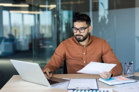 Businessman sitting at a desk in a modern office, working on a laptop while holding and reviewing paper documents.
