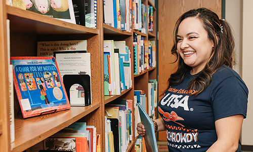 A teacher with a blue UTSA t-shirt on putting a children's book back on the bookshelf.