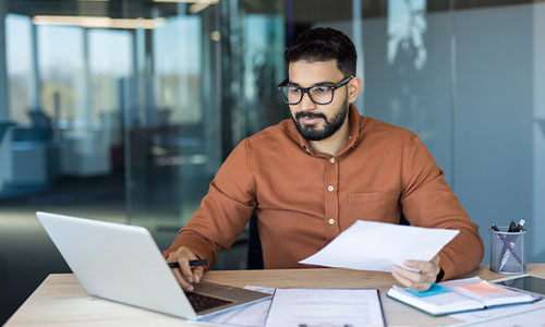 Businessman sitting at a desk in a modern office, working on a laptop while holding and reviewing paper documents.